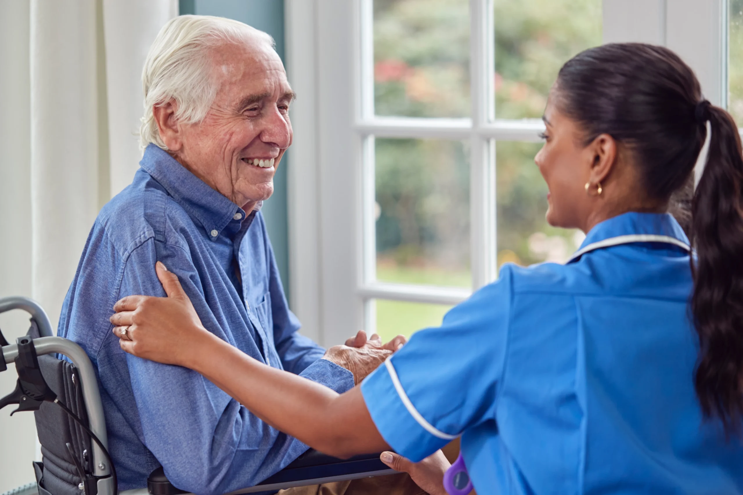 Smiling senior man in a wheelchair interacting with a female caregiver in a blue uniform at home.