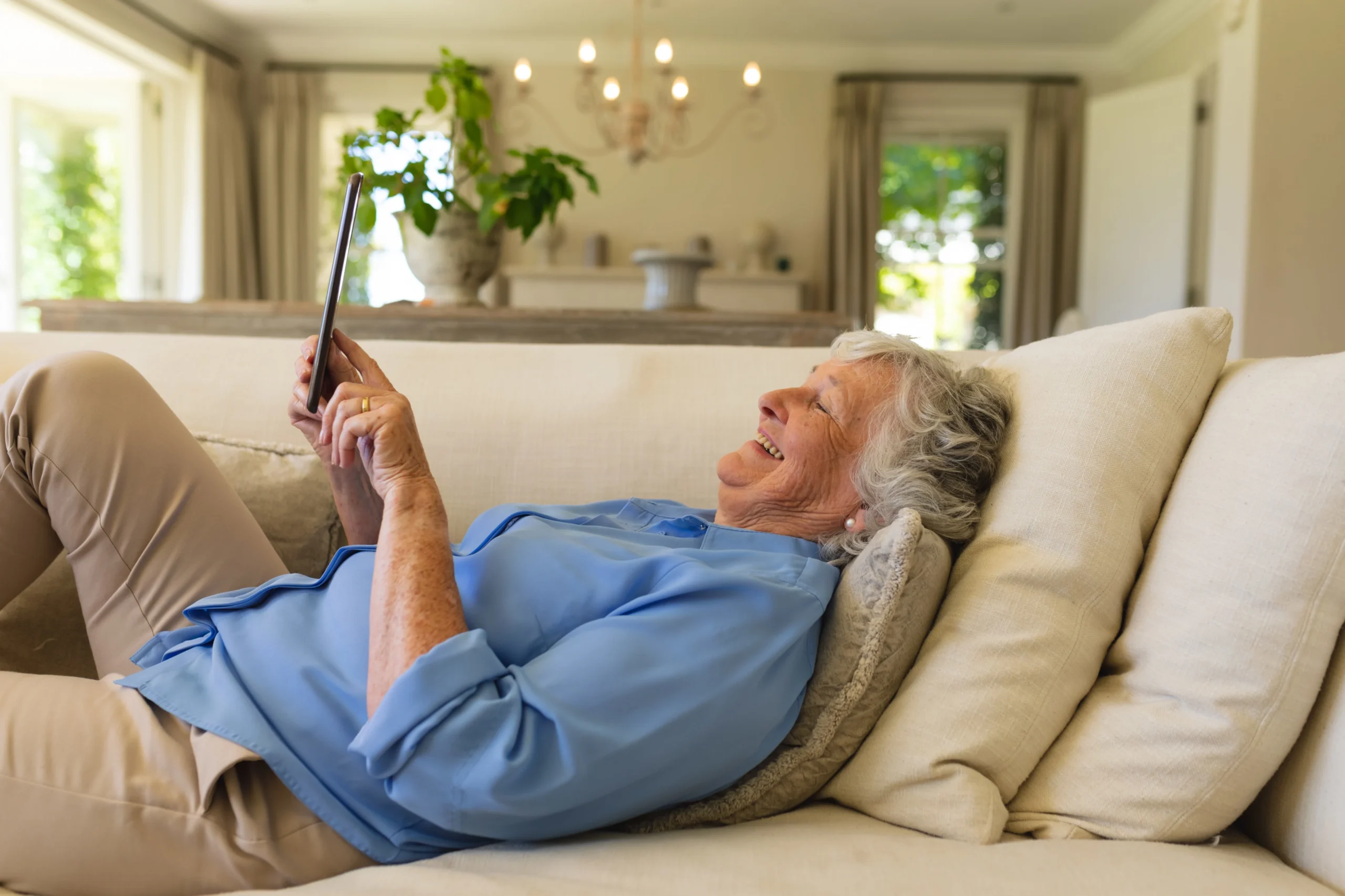 Senior woman relaxing on a sofa and smiling while using a tablet, representing how smart home technology enhances comfort and independence for older adults.